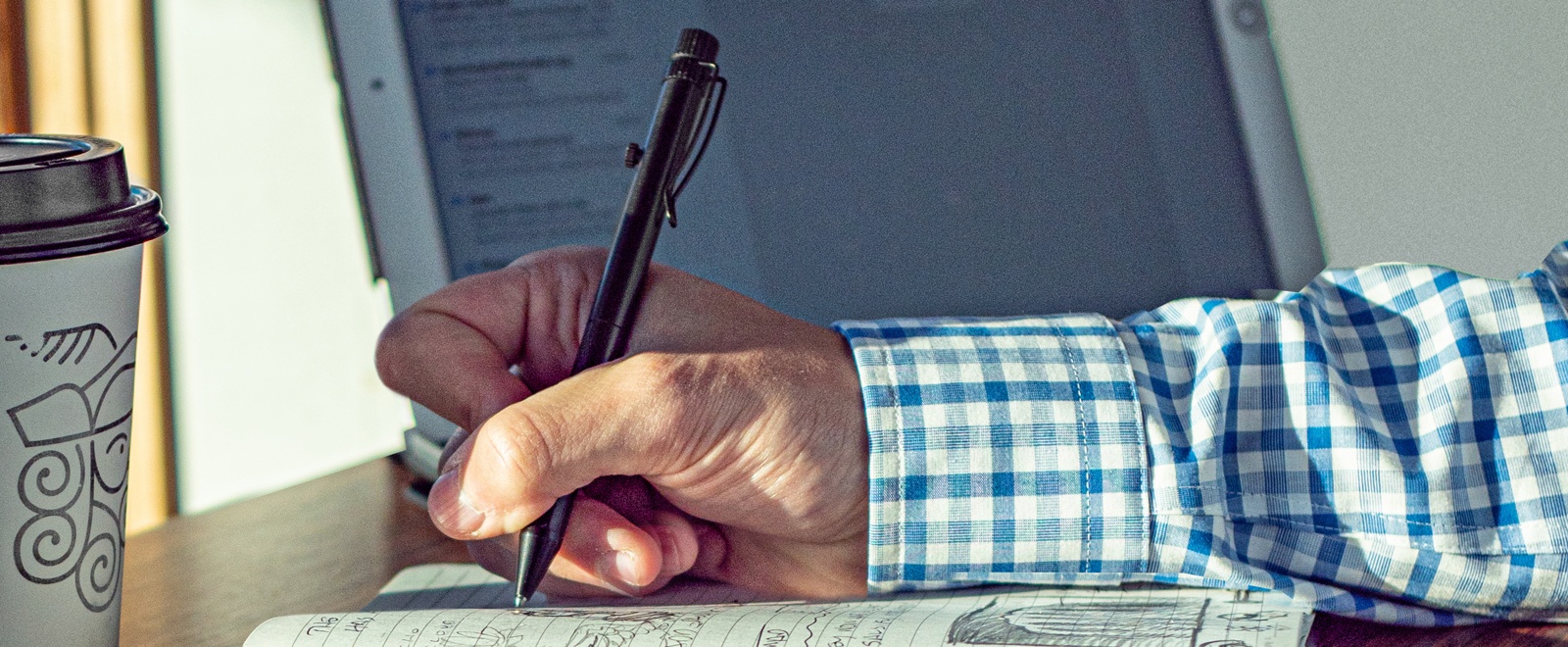 Bolt-action pen in hand writing in a book, computer and coffe cup in the background.